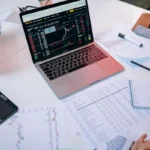 Laptop menampilkan grafik saham dan dokumen analisis keuangan di meja kerja