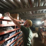 Seorang pekerja gudang sedang menyusun dan mengambil barang dari rak penyimpanan, menggambarkan tugas picker gudang dalam operasional warehouse